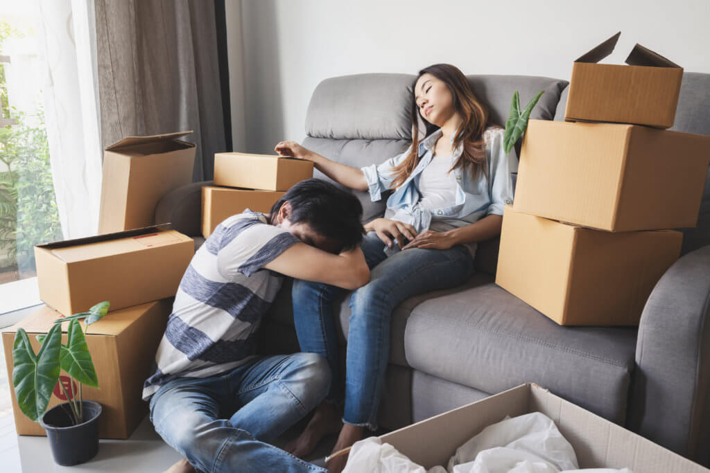 Young tried couple moving in new home, sitting and relaxing on sofa with cardboard boxes