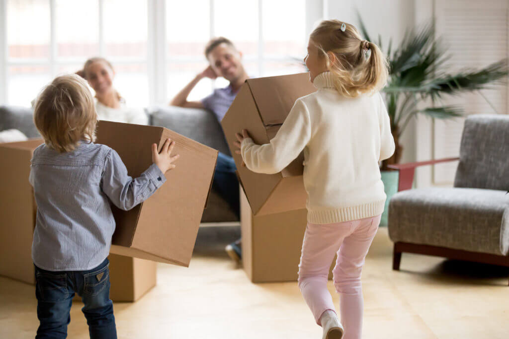 Rear view at kids playing with boxes on moving day, small brother and sister running holding belongings packing unpacking in new home, happy playful children having fun together enjoying relocation