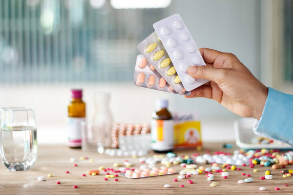 Hand holding medicines pill pack with colorful drugs spread on wooden table in the room background