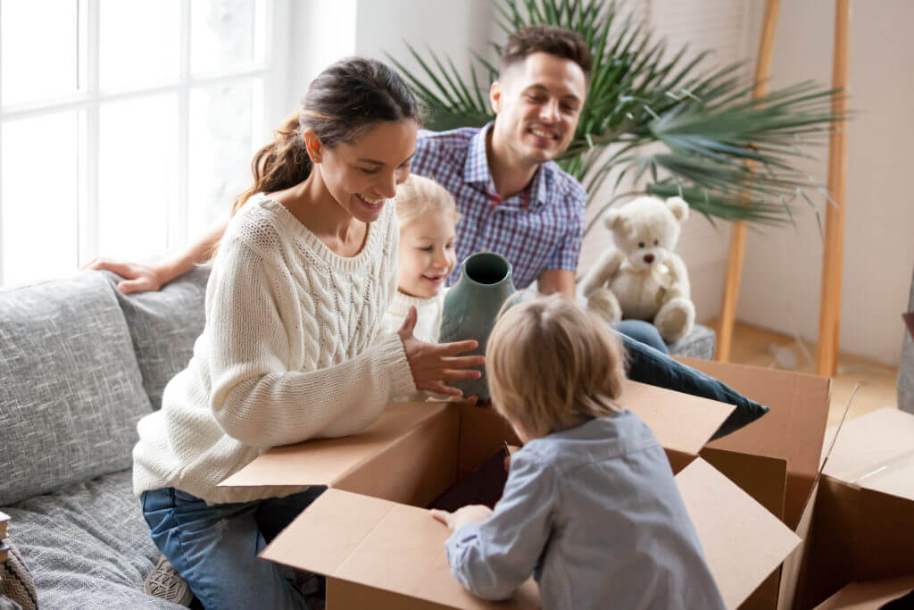 Happy family with two kids unpacking boxes after relocation moving into or settling in new home concept, excited small children helping parents with belongings sitting on sofa in living room together